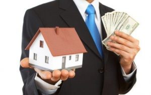 a man in a suit holding a toy house in one hand and fanning out a bundle of cash in his other hand